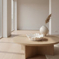 A minimalist interior scene with a round light wood coffee table in a bright, modern room. On the table sits a white scalloped bowl, and a textured white vase with dried grass stands nearby.